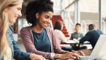 Two female college students sitting in common area on campus looking at laptop.