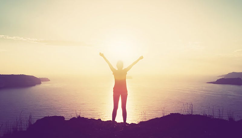 Happy woman with hands up standing on cliff over sea and islands at sunset. Vintage mood, concepts of winner, freedom, happiness etc.