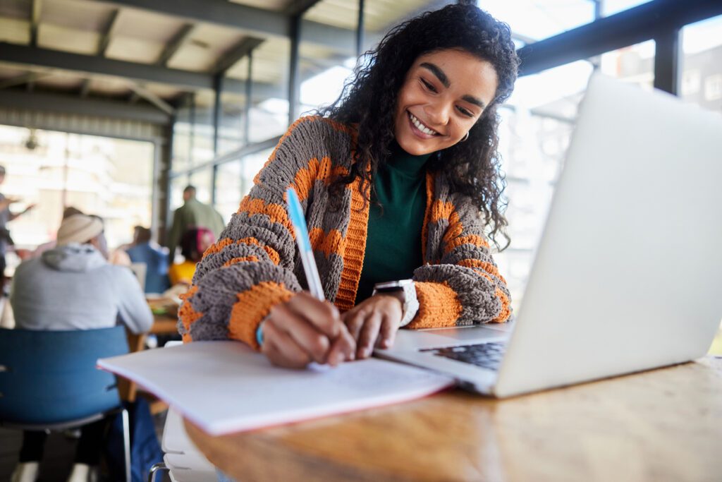 Low angle view of a smiling young female college student writing notes and working on a laptop while doing her homework in a busy campus cafeteria
