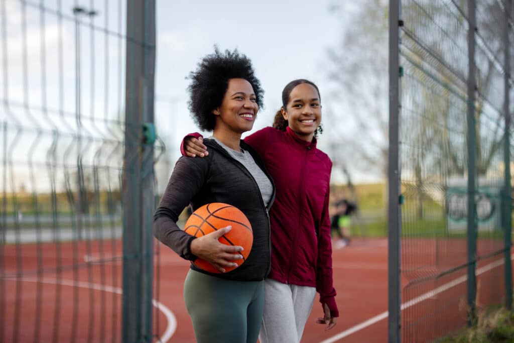African american mother and teenage daughter with a ball on the basketball court outdoors. Sport, people and healthy lifestyle concept