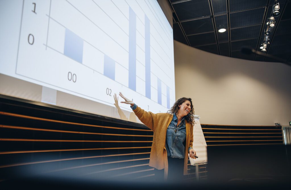 Businesswoman gesturing at projection screen and making a presentation during seminar. Female speaker giving presentation in conference.