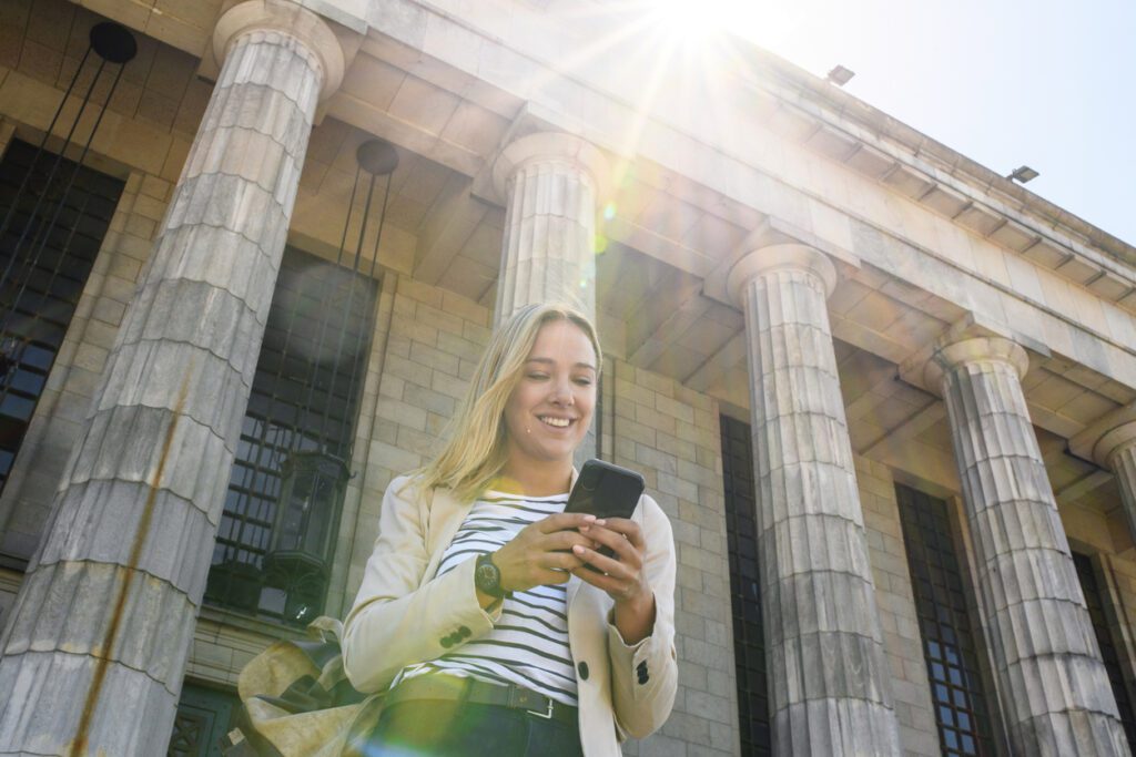Low angle view with lens flare of cheerful female law student in late 20s carrying backpack and checking smart phone as she leaves school.
