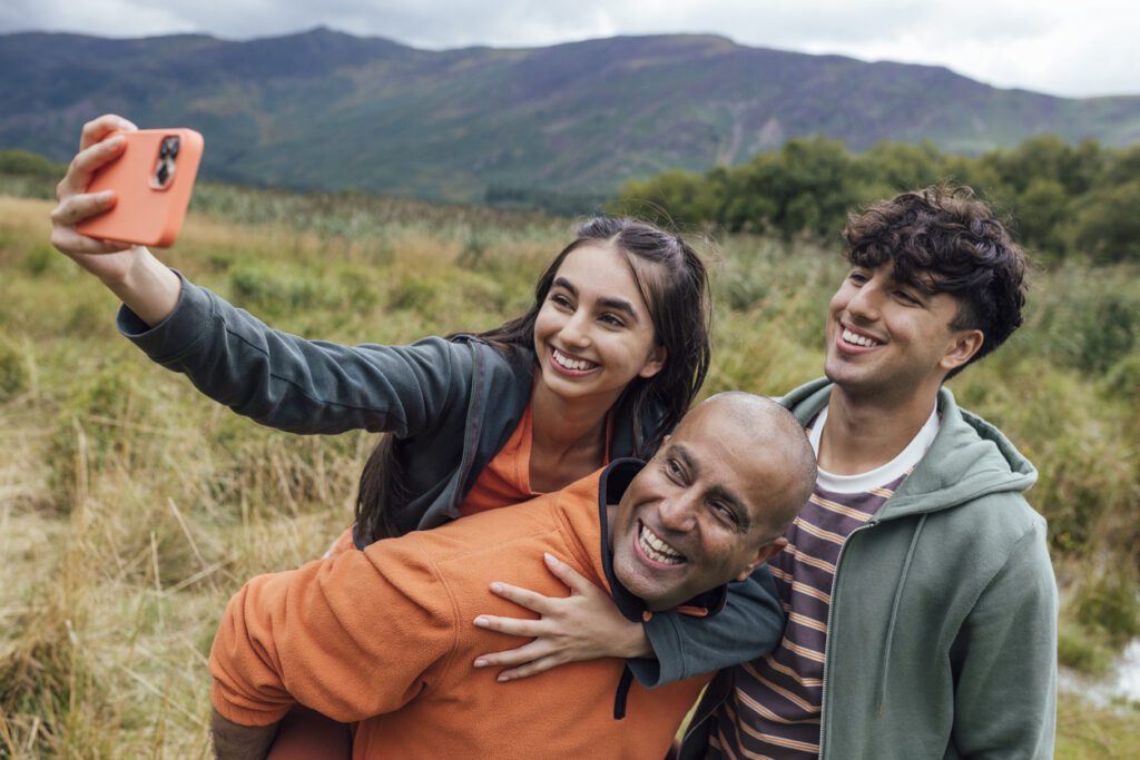 Father, daughter and son on a walk together through the countryside in Keswick, The Lakes. They are using a smart phone to take a selfie while the father gives his daughter a piggyback.