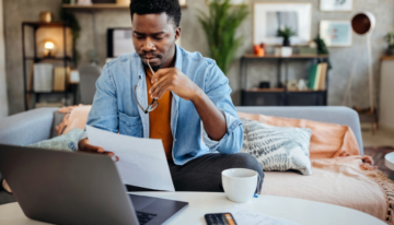 Black man looking over paperwork in front of his computer.