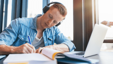 Young man studying in college library with books and laptop.