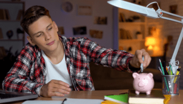 Teenage male student putting money in his piggy bank.