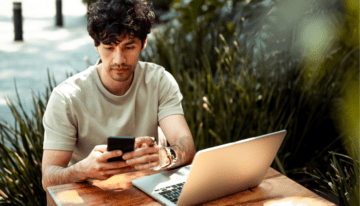 Young man sitting outside at a table with his laptop and looking at his phone.