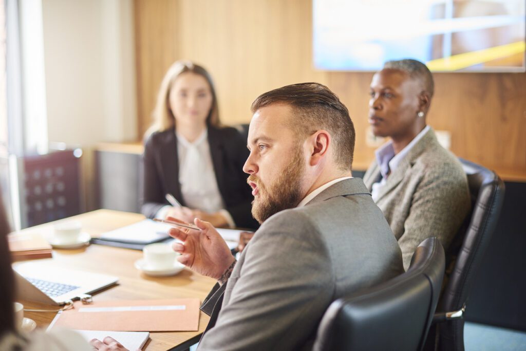 Management team meeting in the boardroom. The team are sat round the conference table discussing the matters at hand as one man asks the others a question