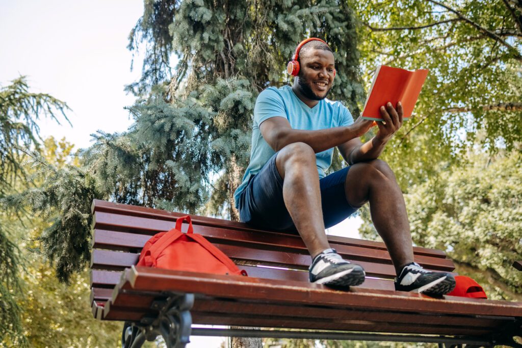 Young mant enjoying and relaxing alone in public park at weekend sitting on park bench listening music using wireless headphones