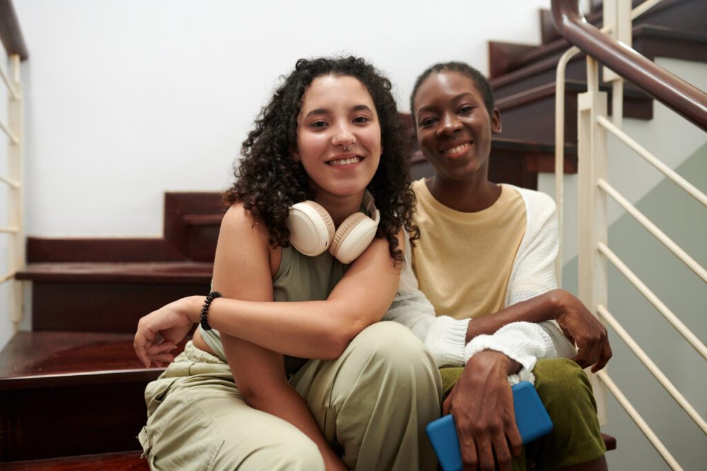 Joyful teenage friends sitting on steps in rented house and smiling at camera