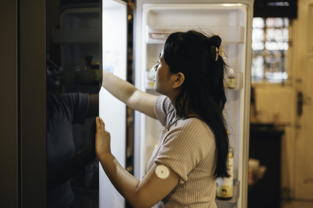 Cropped shot of an attractive young woman using continuous glucose monitor looking in her fridge for a midnight snack at home