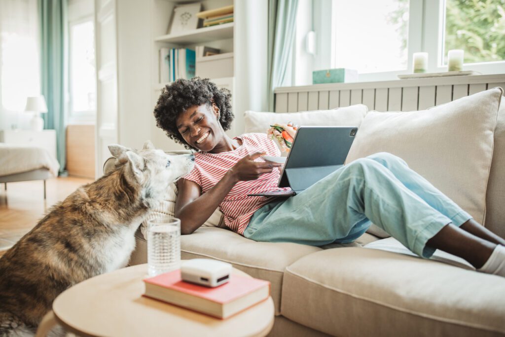 Young woman sitting on sofa using digital tablet and eating sandwich at home with dog