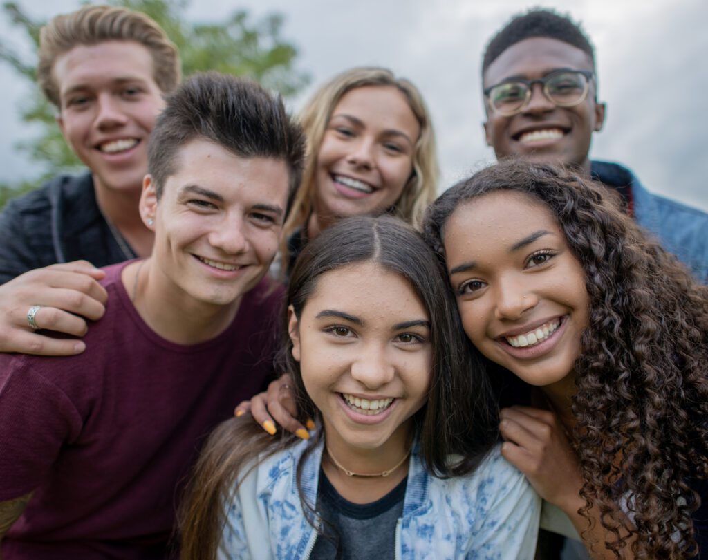 A large group of teens huddle in closely together as they casually pose for a portrait. They are each dressed casually and are smiling as they enjoy time together outside.