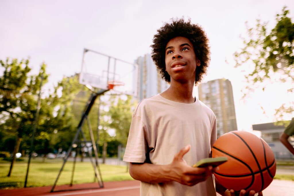Young, happy African American man smiling while holding a basketball and using his phone on an outdoor court, enjoying the summer day and connecting with friends