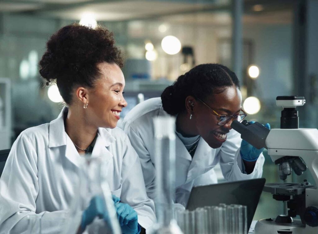 Happy women, microscope and science team research in lab for experiment, analysis and biotechnology. Scientist, collaboration and microbiology test for medical development, healthcare or pharma study