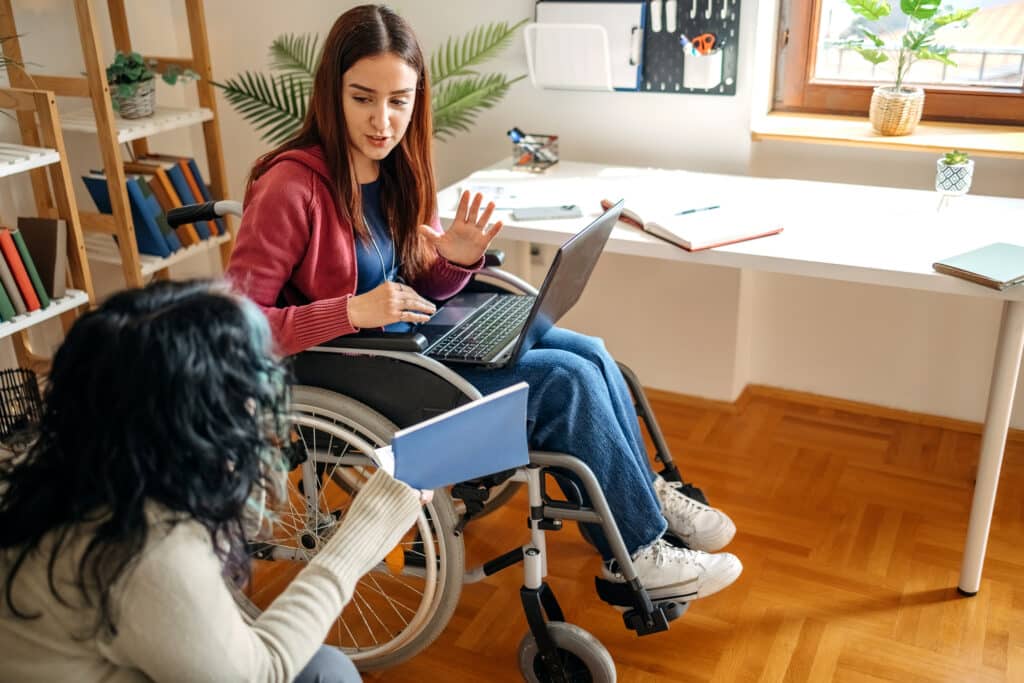 Young female adult with disability preparing for exam studying together with friend at home and talking while looking at laptop and holding a book in hand