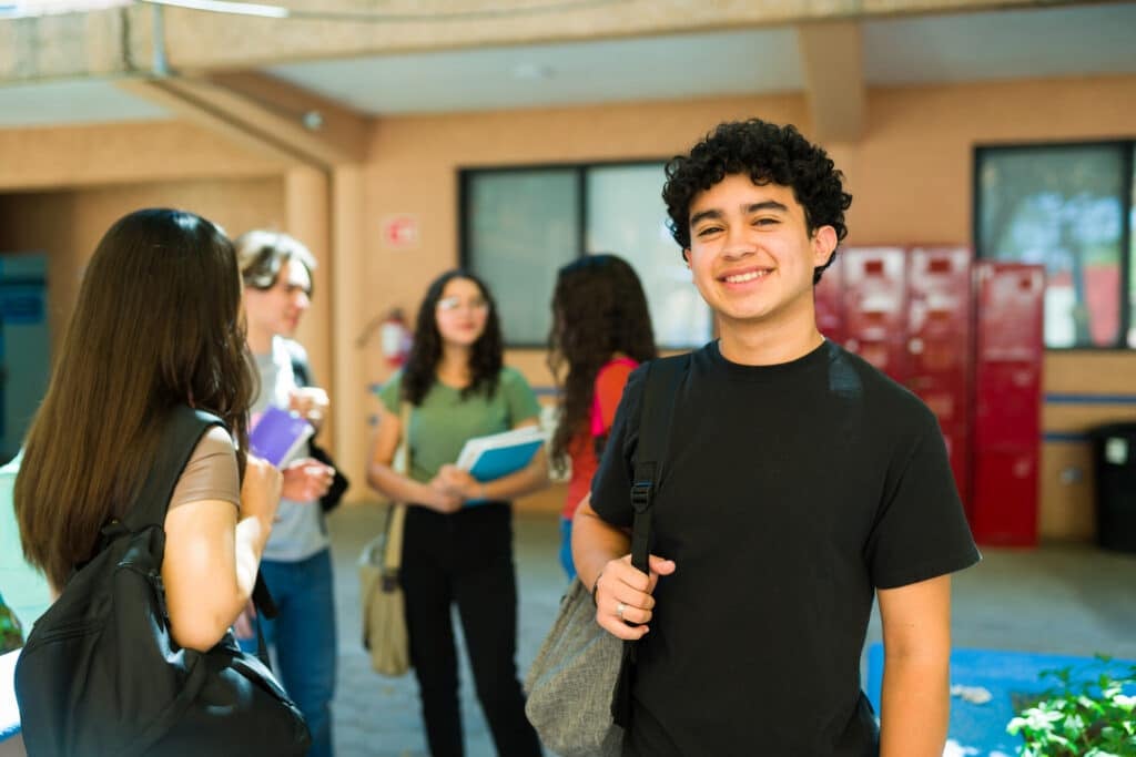 Happy latino high school student smiling in school hallway with classmates chatting in the background