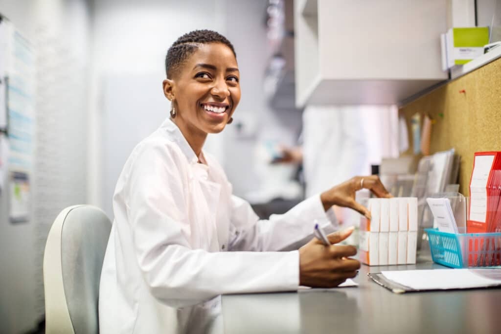 Female chemist sitting at her desk and looking away smiling. African woman pharmacist working at her desk.
