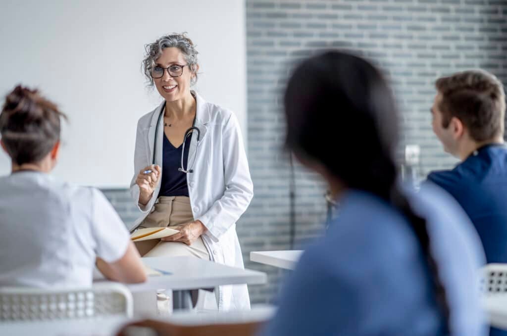 A mature female doctor sits at the front of a classroom as she teaches a small group of students. She has a file folder on her lap as she talks with the students, and is dressed in a white lab coat. The students are seated in front of her and paying close attention as they take notes.