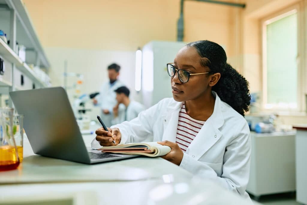 African American female doctor taking notes while working on scientific research in laboratory.