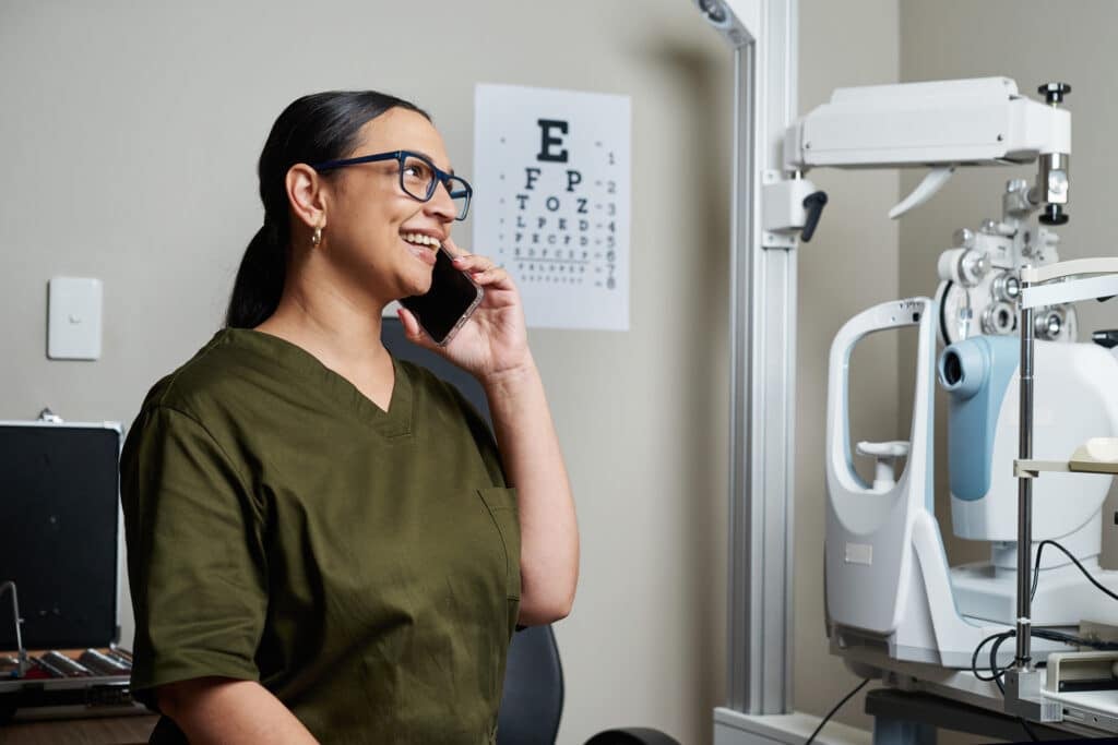 A smiling optometrist and business owner wearing eyeglasses, in an optical clinic setting, confidently talking on the phone. The modern equipment and eye chart in the background emphasize her professional environment.