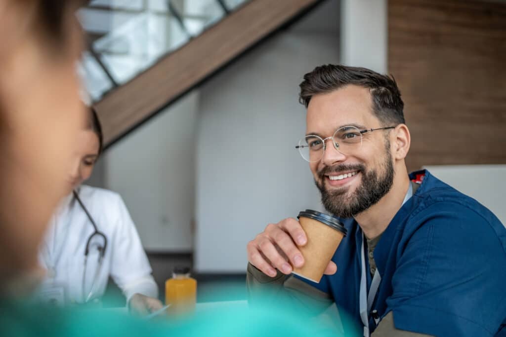 Male healthcare professional smiling and holding a coffee cup, taking a break with colleagues in a modern hospital or clinic environment after a successful meeting