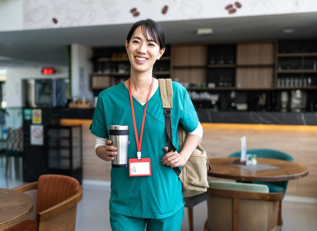 Smiling female nurse wearing green scrubs, holding a reusable coffee cup and a backpack, walking through a modern hospital cafeteria, ready for her shift or break